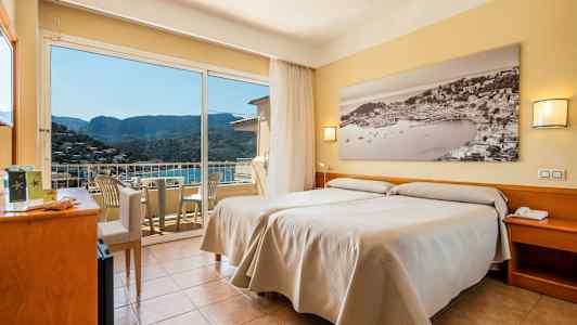 Ett hotellrum med två enkelsängar, ett stort fönster med utsikt över en bergskulle och en balkong med stolar. (AI-genererad text)