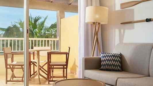 En interiör med soffa och kudde i förgrunden samt utsikt mot en möblerad balkong. (AI-genererad text)