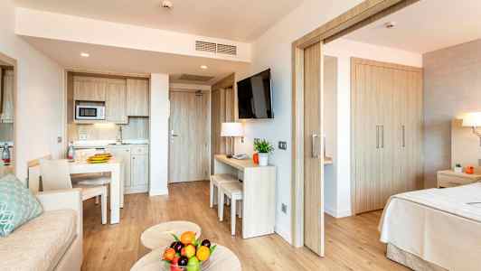 Modernt hotellrum med köksdel, bord, soffa och sovrum. På soffbordet står en skål med färska frukter. (AI-genererad text)