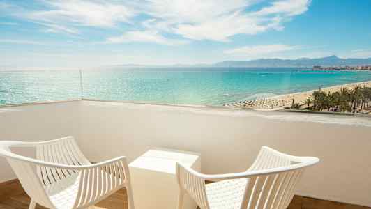 Två vita stolar och ett bord på en balkong med utsikt över havet och stranden. (AI-genererad text)