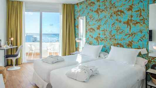 Ett hotellrum med två vita sängar och balkongdörr som leder till en terrass med utsikt mot havet. (AI-genererad text)