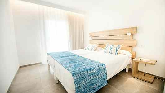 Två enkelsängar med vita lakan och blå överkast, placerade i ett ljust och modernt rum med ett träbord bredvid. (AI-genererad text)