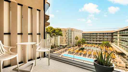 Utsikt från en balkong med två stolar och ett bord, med vy över en pool och hotellbyggnader. (AI-genererad text)