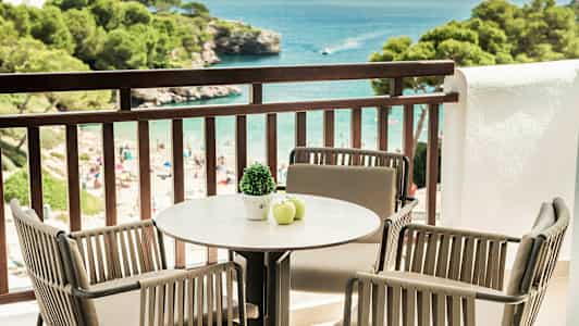 Balkong med bord och stolar som vetter mot havet, stranden och grönskande träd. (AI-genererad text)