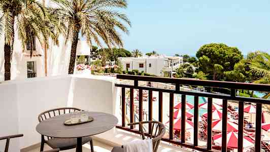 Utsikt från en balkong med stolar och bord mot en pool och omgivande trädgårdar. (AI-genererad text)