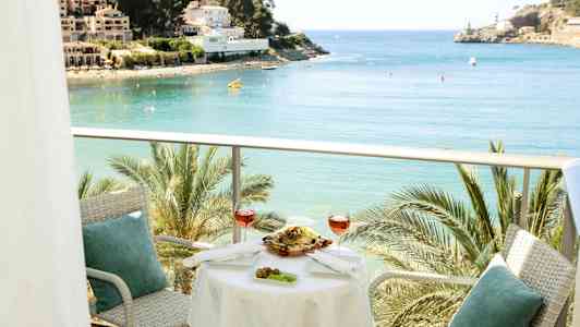 En balkong med bord och stolar, med utsikt över en vik med palmer och byggnader i bakgrunden. (AI-genererad text)