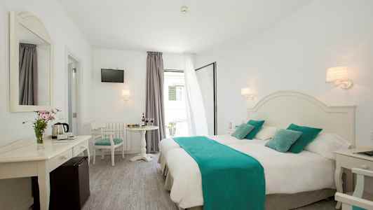 Ett ljust hotellrum med dubbelsäng täckt med vitt sänglinne och turkosa detaljer, bord med stolar samt en spegel och TV. (AI-genererad text)