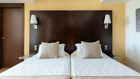 Två enkelsängar med vita lakan och beiga kuddar mot en mörkbrun sänggavel, med vägglampor ovanför. (AI-genererad text)