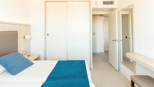 Ett ljust hotellrum med en säng med blå sängkläder, ett nattduksbord och garderobsdörrar i bakgrunden. (AI-genererad text)