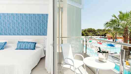 Ett hotellrum med två sängar, en blå sänggavel och tillgång till en balkong med utsikt över en pool och trädgård. (AI-genererad text)