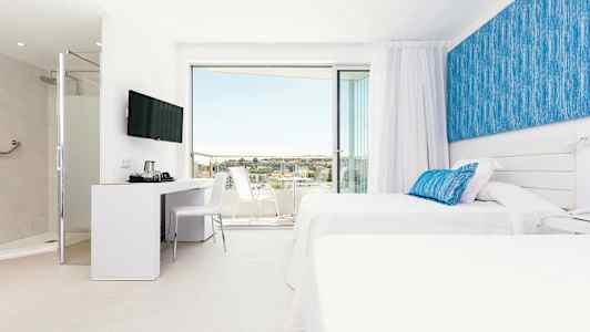 Ett modernt hotellrum med två vita sängar och blå dekorationer. Rummet har en sittgrupp och en inglasad balkong med utsikt. (AI-genererad text)