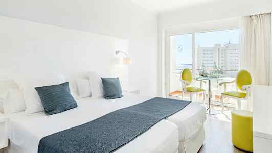 Ett ljust hotellrum med två enkelsängar, vita möbler och en sittgrupp nära en glasdörr som leder till en balkong. (AI-genererad text)