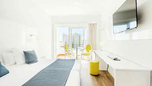 Ljust hotellrum med dubbelsäng, TV på väggen och sittmöbler vid fönster. (AI-genererad text)