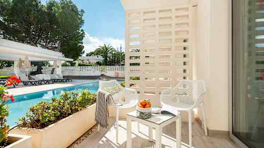 Bild av en balkong med två vita stolar och bord med apelsiner, med utsikt över en pool och trädgård. (AI-genererad text)