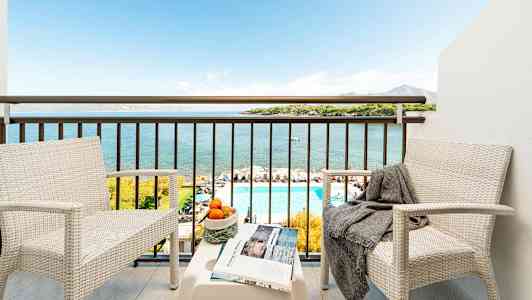 Två vita stolar med ett bord på en balkong som erbjuder utsikt över havet, en pool och landskap. (AI-genererad text)
