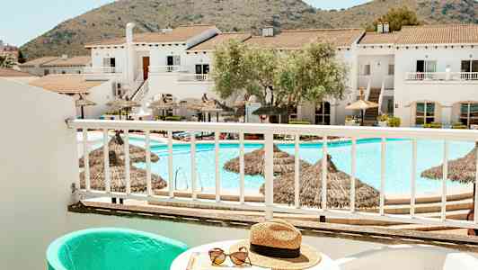 Utsikt från en balkong mot en pool och byggnader med berg i bakgrunden. (AI-genererad text)