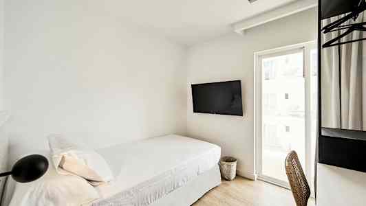 Ett hotellrum med en enkel säng, väggmonterad TV, och ett skrivbord med stol bredvid ett fönster. (AI-genererad text)