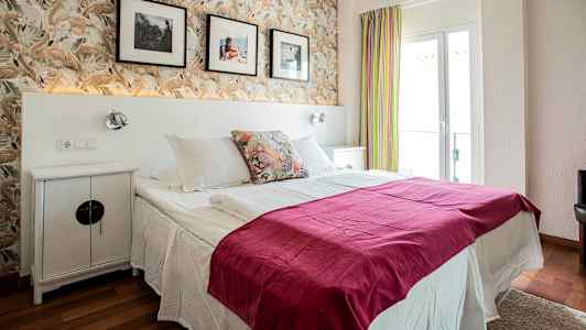 Ett hotellrum med en dubbelsäng, dekorerad med rosa sängöverkast och huvudkuddar. Bakom sängen finns en tapet med fjädermönster och väggdekorationer. (AI-genererad text)
