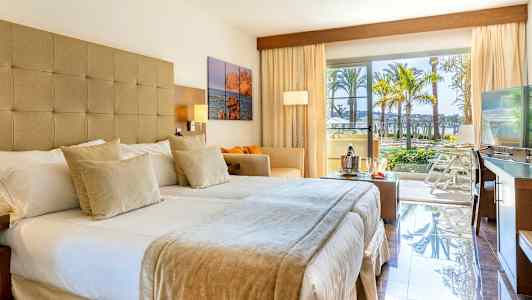 Hotellrum med dubbelsäng, soffa, TV och stora fönster som öppnar ut mot en balkong med utsikt över palmer och poolområde. (AI-genererad text)