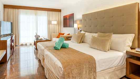 Hotellrum med en stor säng, sittgrupp och ljusa gardiner framför en balkong. (AI-genererad text)