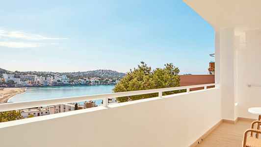 Utsikt från en balkong över en strand vid en vik, med byggnader och grönska i bakgrunden. (AI-genererad text)