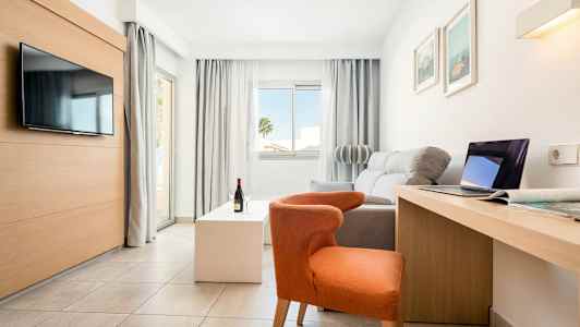 Ljust, modernt hotellrum med soffa, stol, TV, skrivbord och stora fönster. (AI-genererad text)