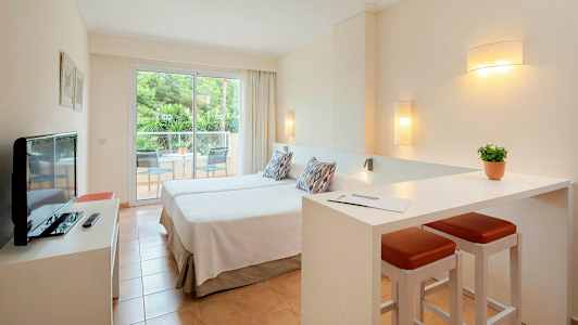 Ett hotellrum med två enkelsängar, en liten sittgrupp och en balkong med utsikt mot gröna träd. (AI-genererad text)