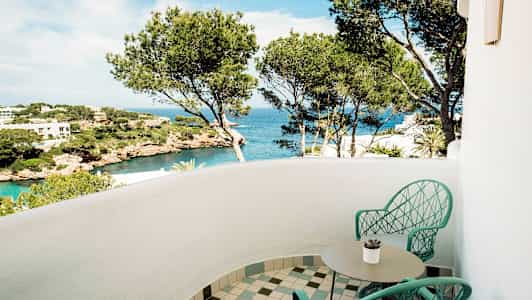 En balkong med två stolar och ett bord, utsikt över havet och landskap med tallar. (AI-genererad text)