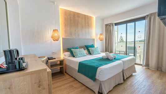 Ett hotellrum med två enkelsängar, en träpanel vid sängen, turkosa lakan och kuddar, samt en balkong med utsikt. (AI-genererad text)