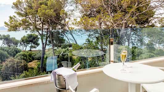 En balkong med utsikt över träd och hav. På balkongen står ett vitt bord med ett glas champagne och en ställning med en kylhink för en champagneflaska. (AI-genererad text)