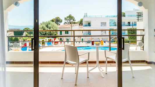 En balkong med två stolar och ett bord med drycker och snacks. Utsikt över ett poolområde och omgivande byggnader. (AI-genererad text)