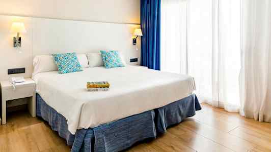 Ett hotellrum med en bred säng, vit sänggavel, blå kuddar och gardiner samt en ljus träparkett. (AI-genererad text)