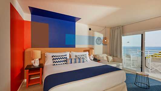 Ett rum med en dubbelsäng, dekorativt målade väggar, sittmöbler och en utsikt mot havet genom stora fönster. (AI-genererad text)