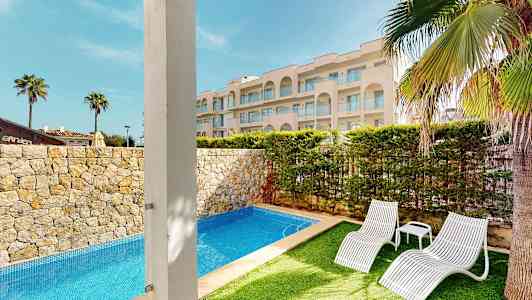 Bild av en utomhuspool omgiven av stengärder och grönskande trädgård, med två vita solstolar och ett vackert hotell i bakgrunden. (AI-genererad text)