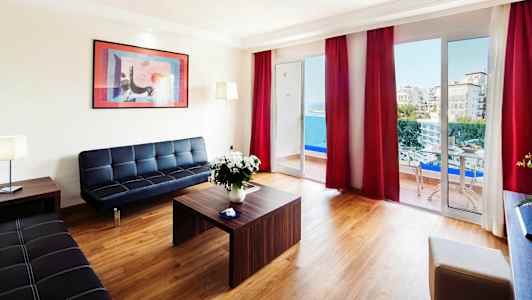 Ett modernt vardagsrum med utsikt mot havet genom stora fönster och balkong, möblerad med svarta soffor och ett träbord. (AI-genererad text)