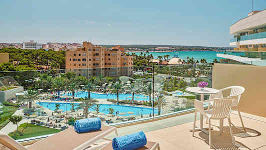 Utsikt från en balkong över en pool och trädgård med palmer, hotellbyggnader och hav i bakgrunden. (AI-genererad text)