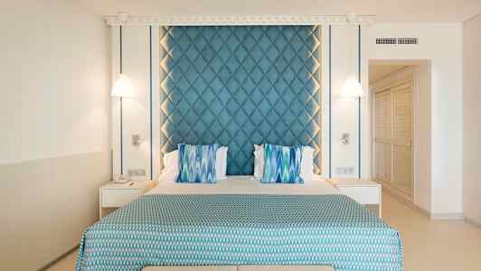 Hotellrum med en dubbelsäng som har blå och grön dekor samt två sänglampor på vardera sida. (AI-genererad text)