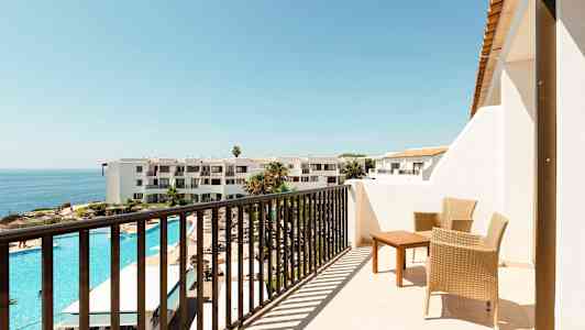 En balkong med två flätade stolar och ett bord, utsikt över ett hotellområde med utomhuspool och havet i bakgrunden. (AI-genererad text)