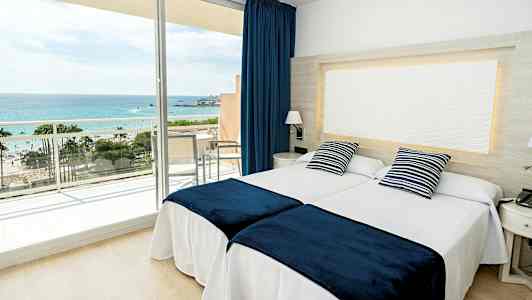 Ett hotellrum med två sängar, blåvita prydnadskuddar och blå överkast, samt en balkong med utsikt mot havet. (AI-genererad text)