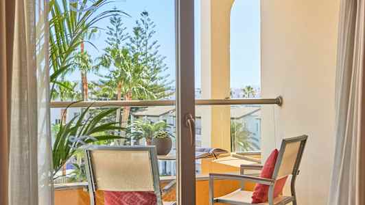 Utsikt genom en balkong med två stolar och ett bord, omgivande växter och byggnader. (AI-genererad text)