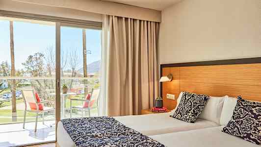 Bild av ett hotellrum med en dubbelsäng, ett bord med stolar på balkongen och utsikt mot palmer utanför. (AI-genererad text)