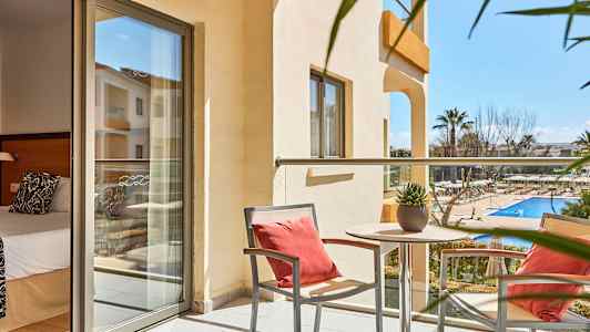 En balkong med två stolar och ett bord med en växt, utsikt över en utomhuspool och omgivande trädgård. Ett rum med säng skymtar i bakgrunden. (AI-genererad text)