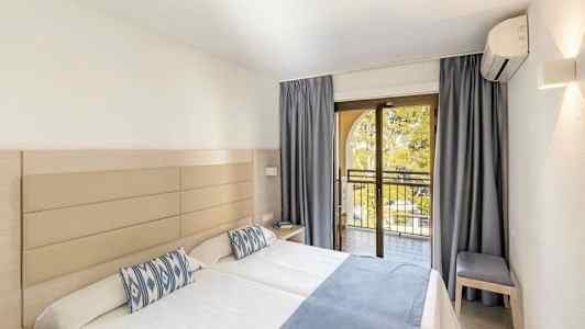 Ett hotellrum med två enkelsängar, ljus inredning, blåa sängöverdrag och kuddar, samt en balkong med öppna glasdörrar. (AI-genererad text)