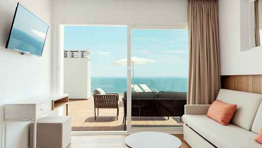 Ett ljust rum med en soffa och bord, som har en utomhusbalkong med sittmöbler och utsikt över havet. (AI-genererad text)