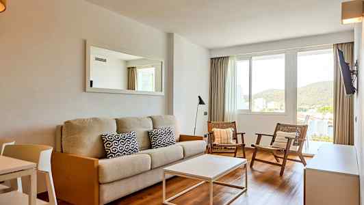 Ett vardagsrum med en beige soffa, två stolar och ett vitt soffbord. Det finns ett fönster med utsikt mot grönska. (AI-genererad text)