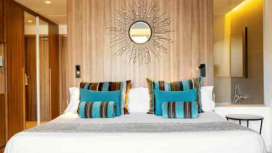 Ett hotellrum med en stor säng dekorerad med färgglada kuddar i blått och randigt mönster, en trävägg med ett spegelmotiv och modern belysning. (AI-genererad text)