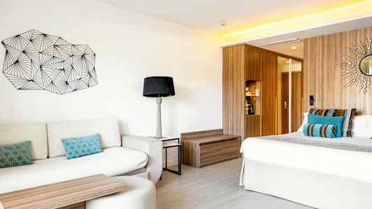 Ett modernt hotellrum med en soffa, färgglada kuddar, ett soffbord, en säng med dekorativa kuddar samt en träpanel bakom. På väggen finns konst och ett litet runt spegeldekoration. (AI-genererad text)