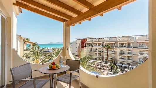 Balkong med två stolar och ett bord med fruktskål, havsutsikt och hotellbyggnader i bakgrunden. (AI-genererad text)