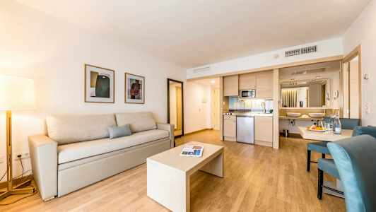 Ett modernt hotellrum med en beige soffa och ett bord på ett trägolv, köksdel och badrum i bakgrunden. (AI-genererad text)