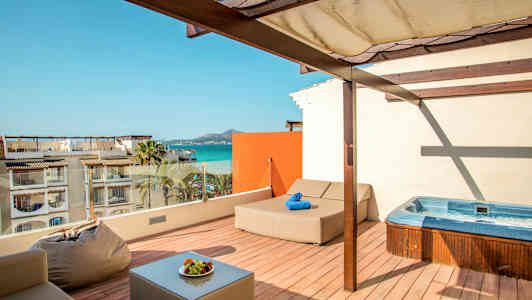 Takterrass med jacuzzi, solbädd och sittplatser, utsikt mot havet och byggnader. (AI-genererad text)